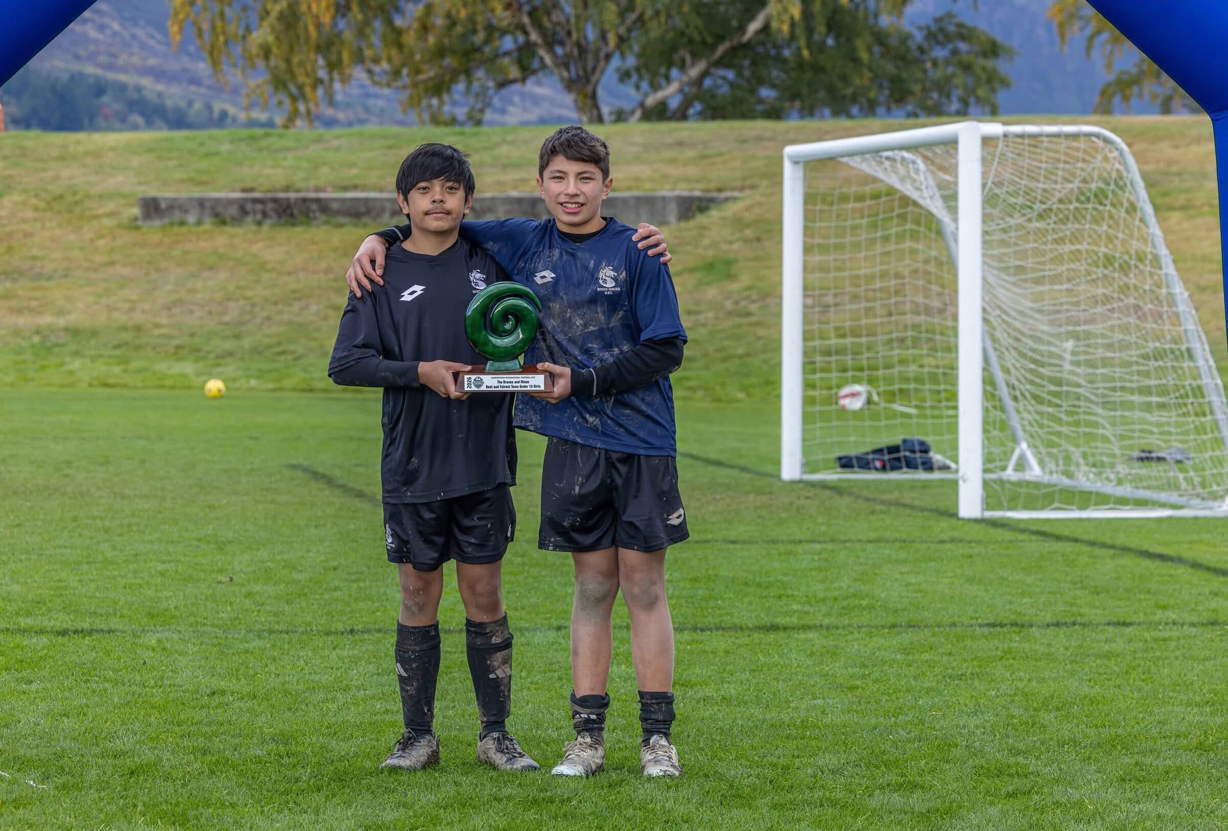 Two young footballers holding a trophy between them beside the goal