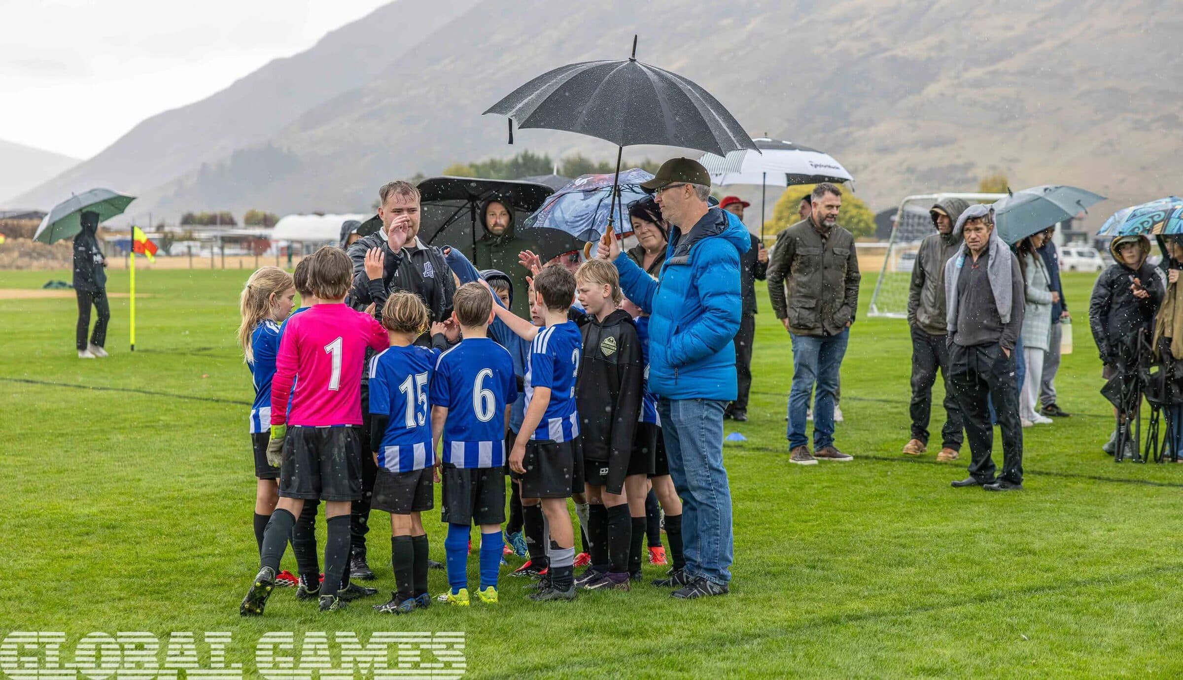 Junior teams lined up on the field with supporters and officials, mountains behind